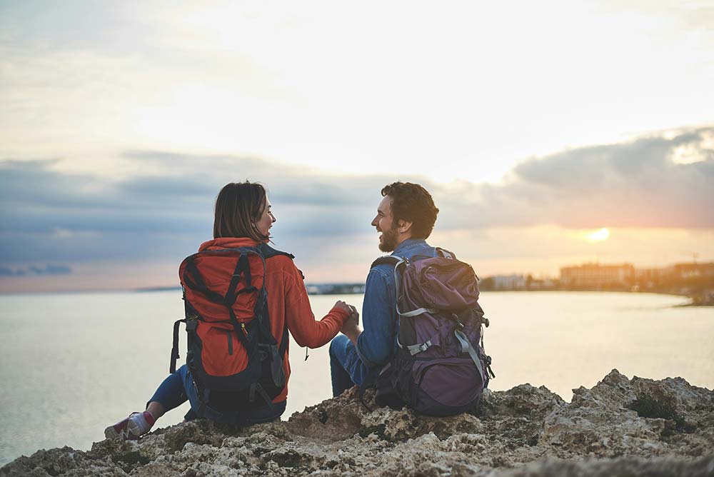 A couple holding hands and smiling on a rocky cliff overlooking the ocean at sunset