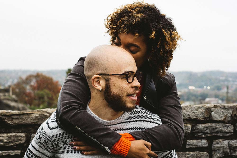 A woman hugs a man from behind and kisses his head.