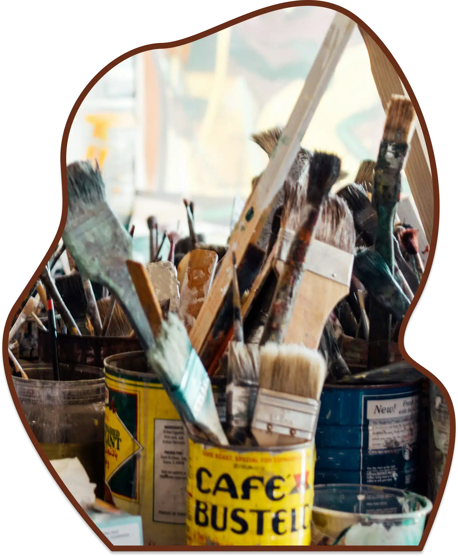 A cluster of well-used paintbrushes sitting in old metal coffee cans inside an artist studio, framed in a wavy border.