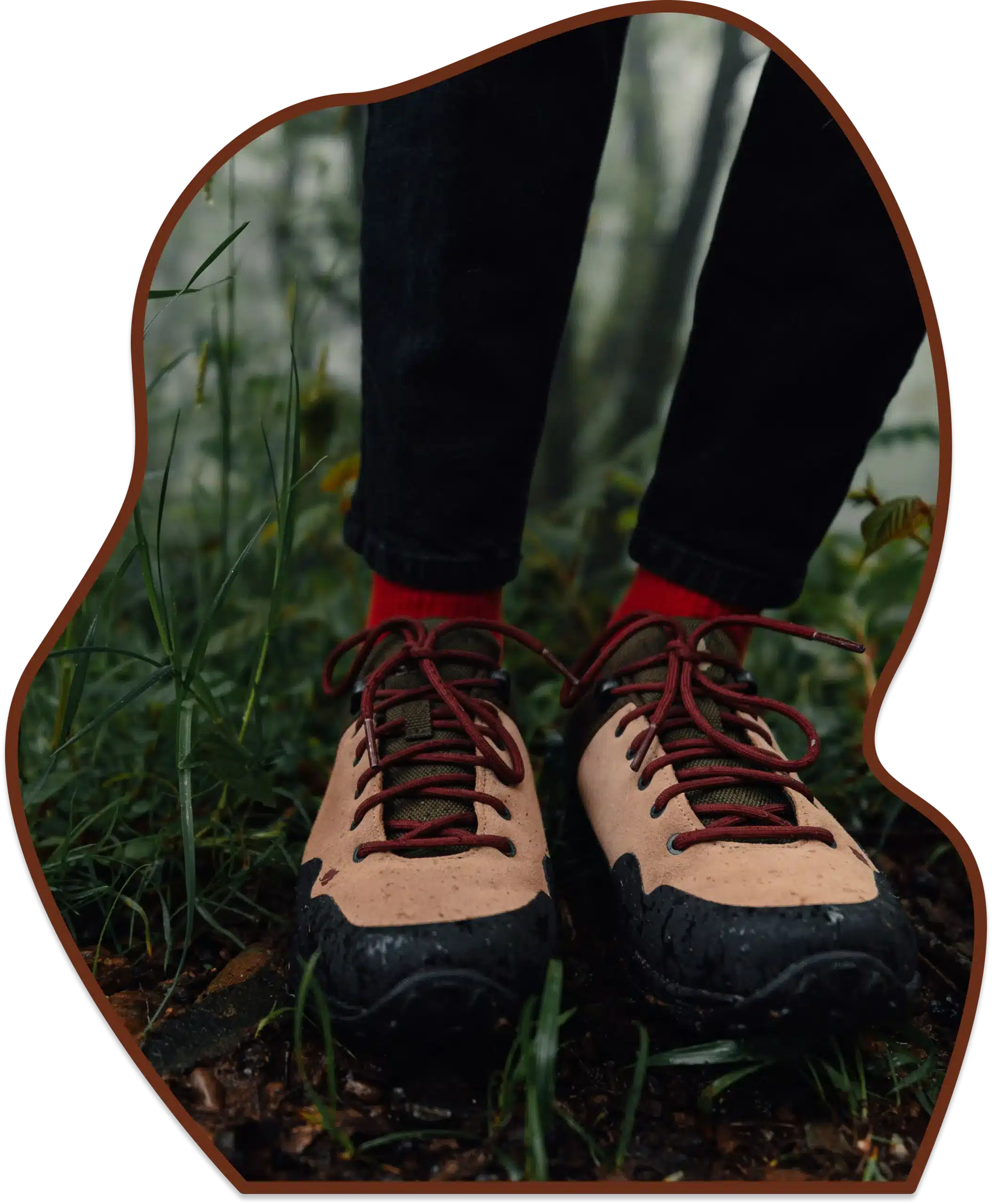 Close-up of a person's feet wearing tan and black hiking shoes with red socks and laces, standing on a wet, muddy forest path surrounded by green grass.