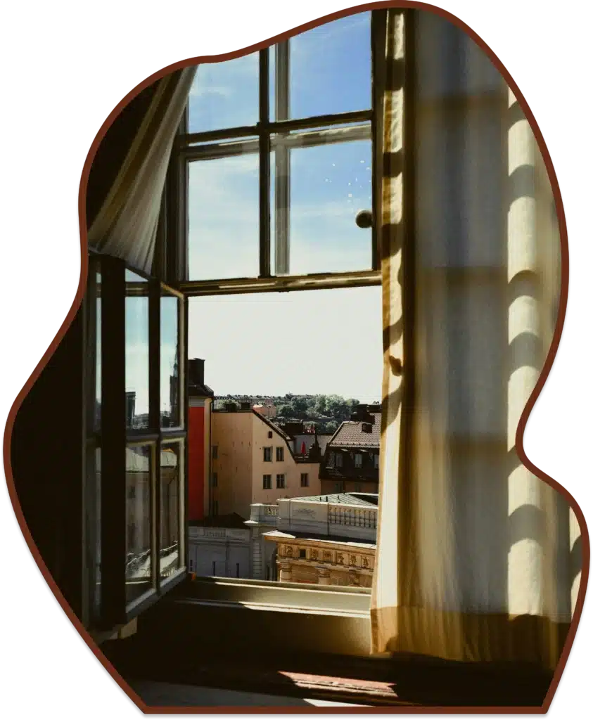 Open window with flowing curtains overlooking sunlit city rooftops, capturing a peaceful interior scene with scenic urban views and natural light.
