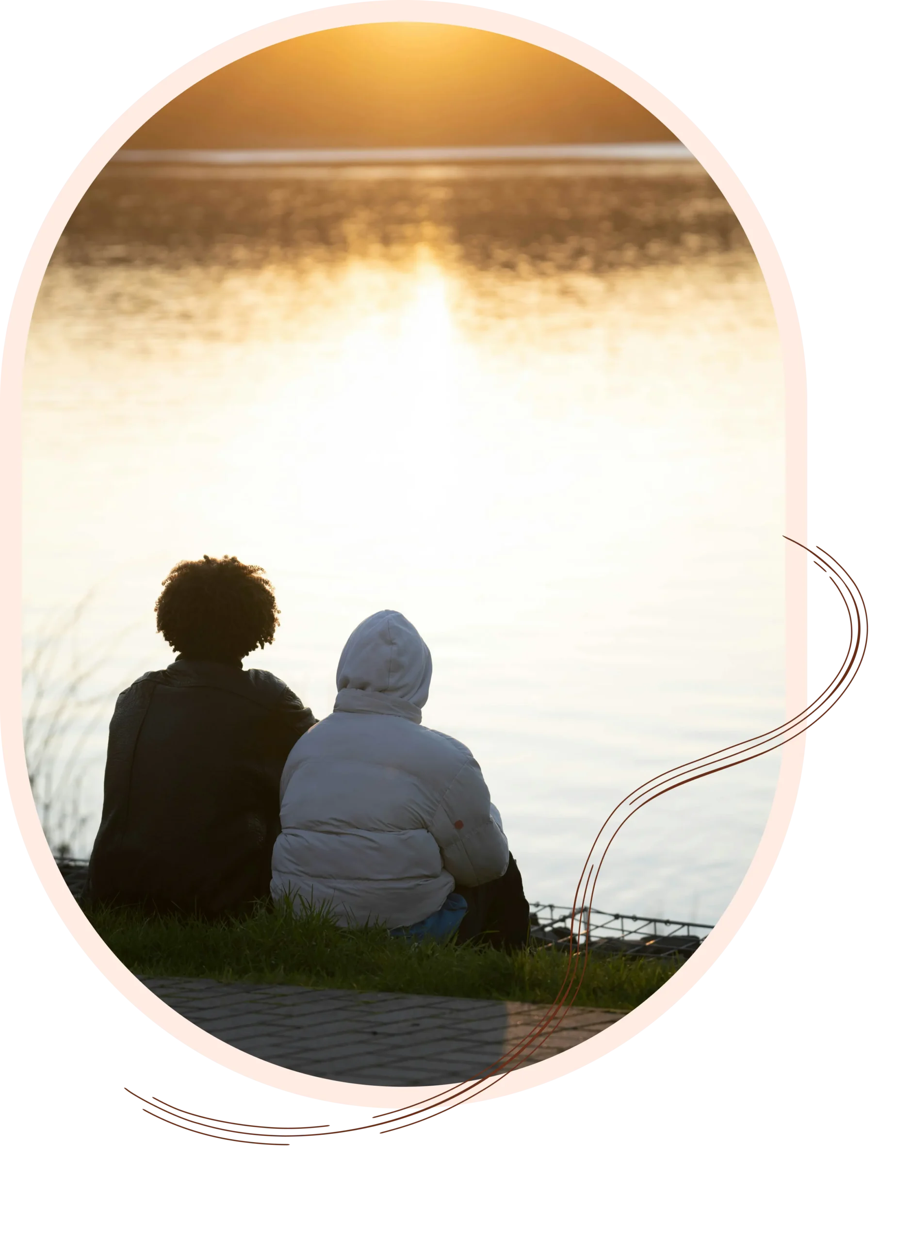 Two people sitting on a grassy bank with their backs to the camera, watching a bright, golden sunset reflect over a calm lake.